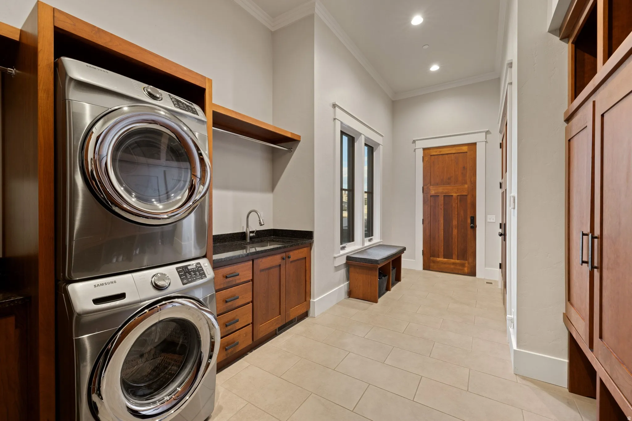 Dedicated laundry space featuring a stacked washer and dryer, wood-finish cabinetry with a sink, stone countertop, and tile flooring