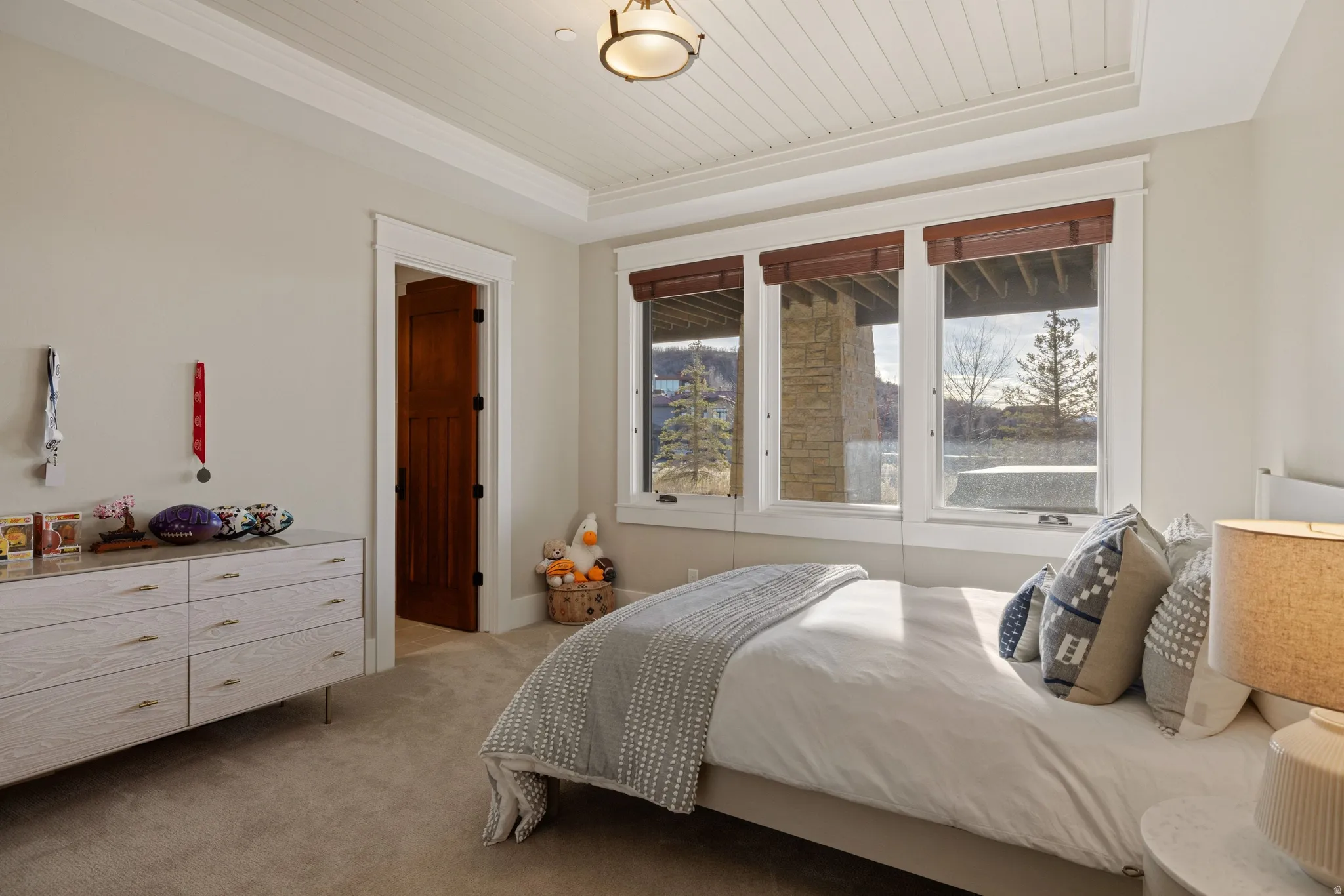 Tray ceiling with beadboard paneling, three large windows with wood-tone blinds, warm beige wall color, and plush carpet flooring