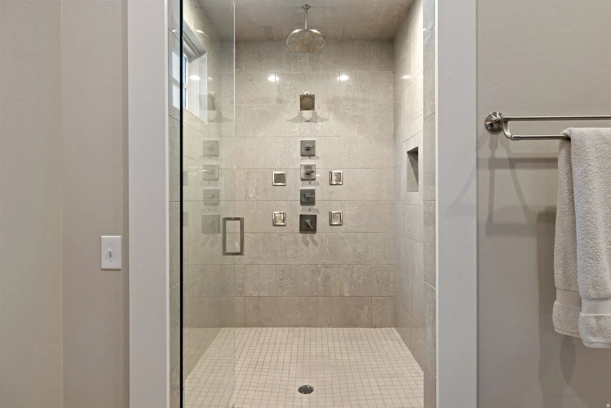 Glass-enclosed shower with a rainfall showerhead, multiple body sprays, recessed niche, and light-toned tile flooring