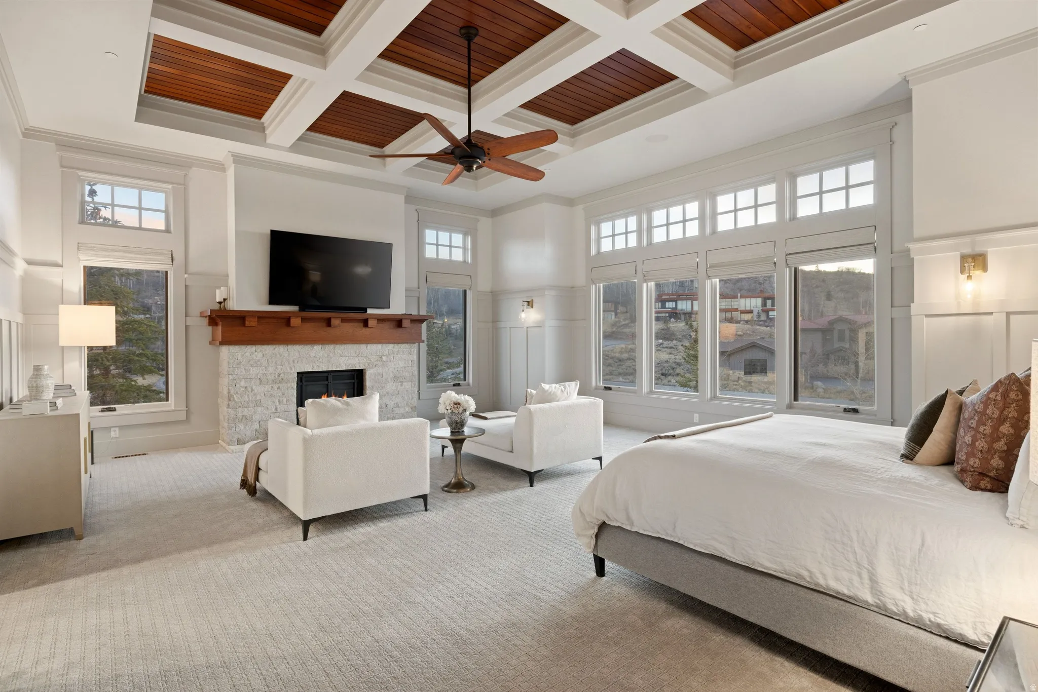 Spacious room featuring a coffered ceiling with wood paneling, a stone-faced fireplace with a wood mantel, extensive window banks, wall paneling, and wall-mounted sconce lighting