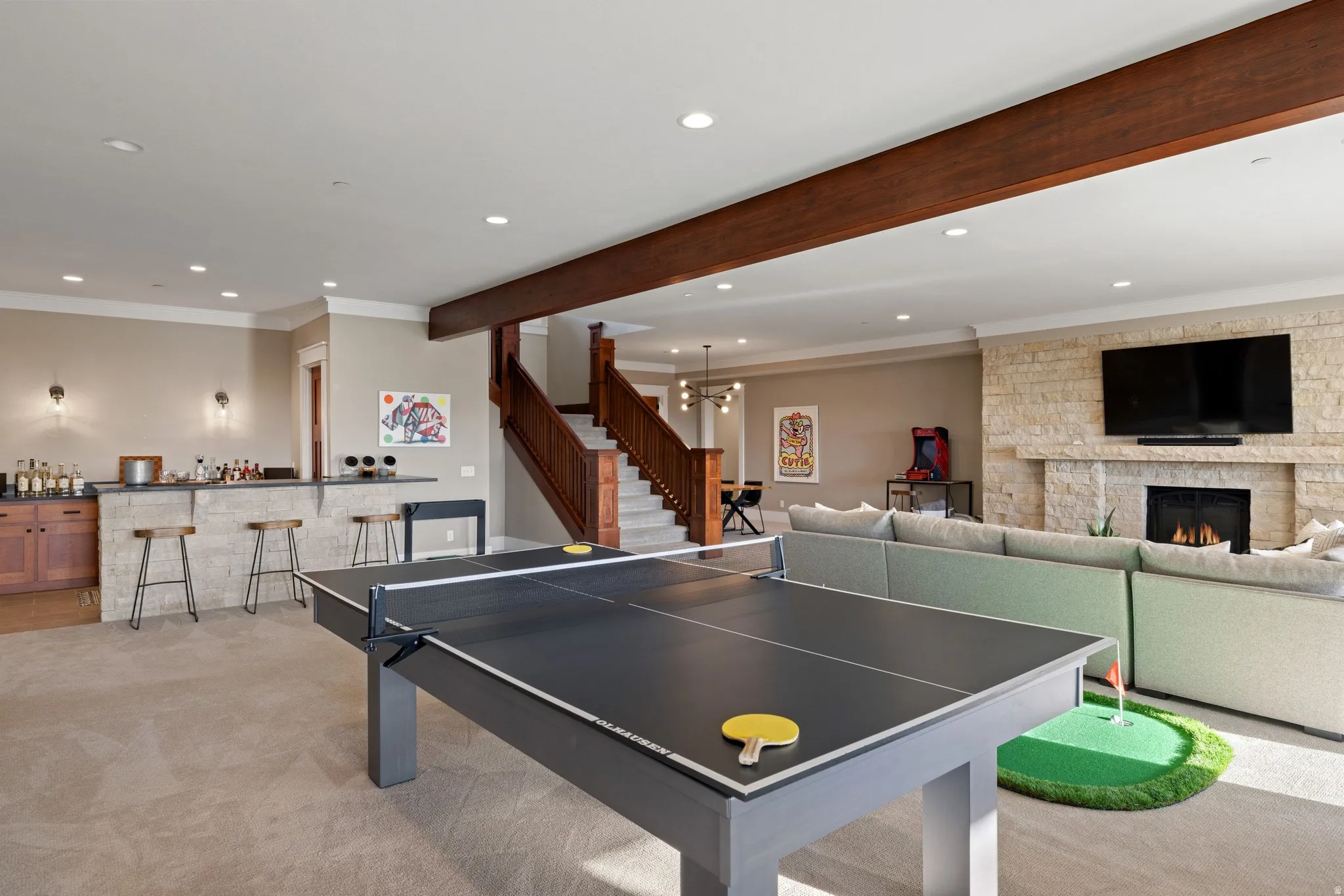 Open-concept recreation area featuring a built-in wet bar with cabinetry, a stone-clad fireplace with mantel, exposed wood ceiling beam, recessed lighting, and a wood staircase with balusters