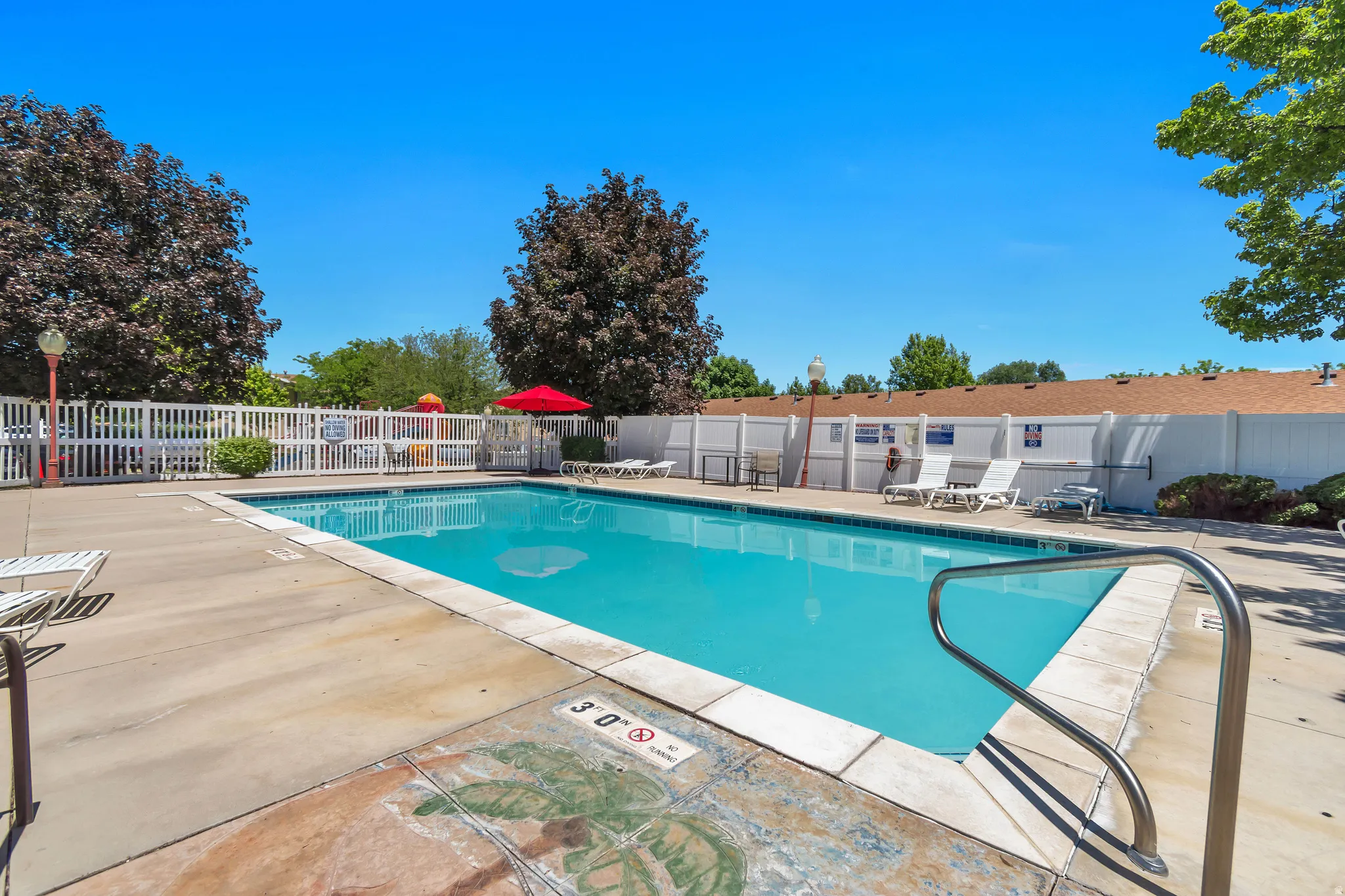 Outdoor swimming pool featuring a white perimeter fence, concrete decking, and mature trees