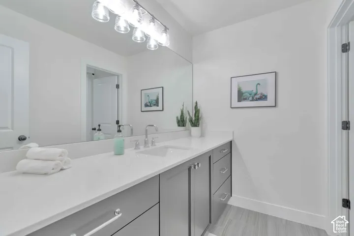 Grey cabinetry with polished hardware, white countertop with an integrated rectangular sink, wall-spanning mirror, multi-bulb vanity light fixture, and tile flooring
