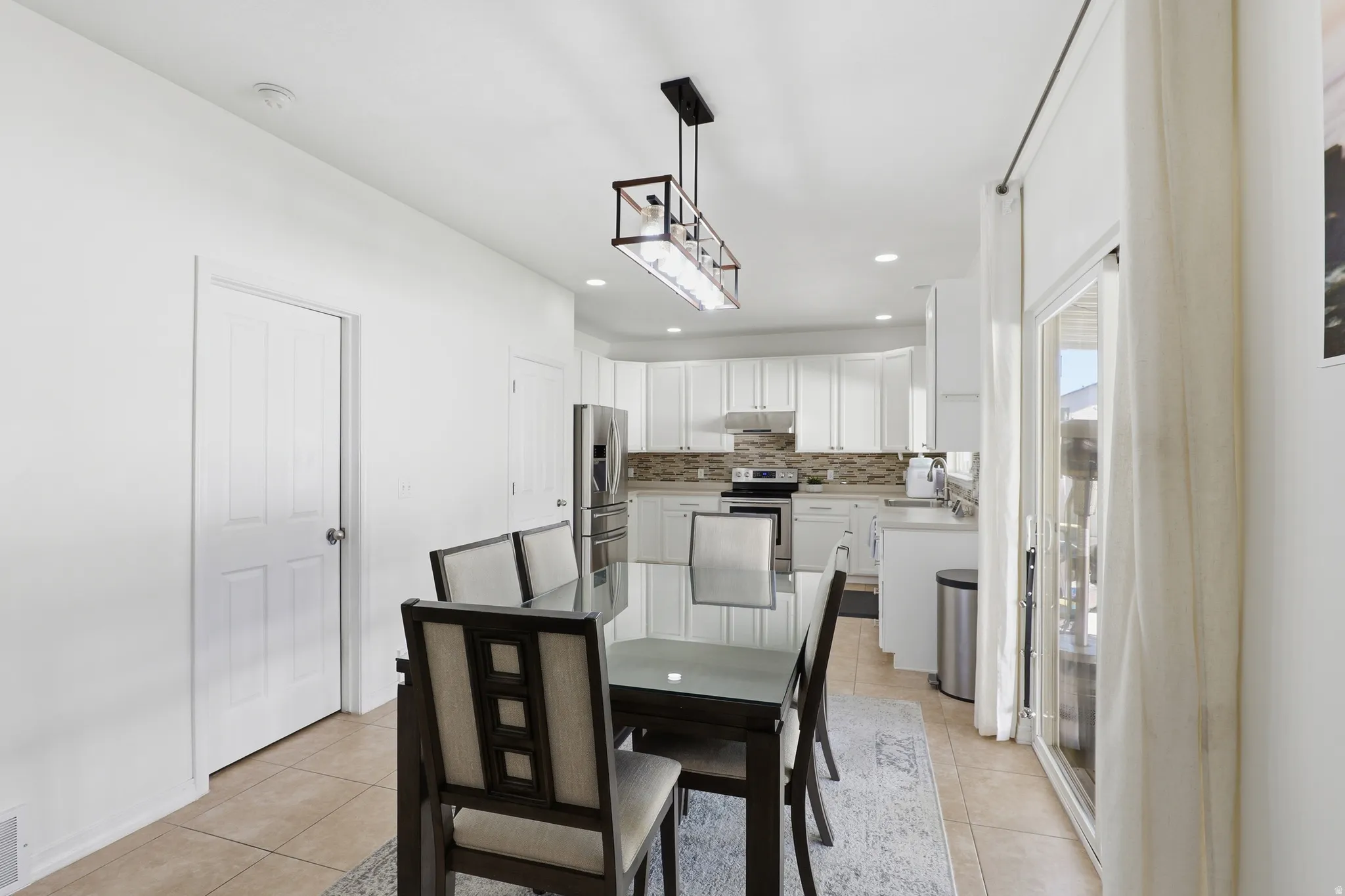 Modern kitchen and dining area featuring recessed lighting, a linear chandelier, ample white cabinetry, stainless steel appliances, and a glass sliding door