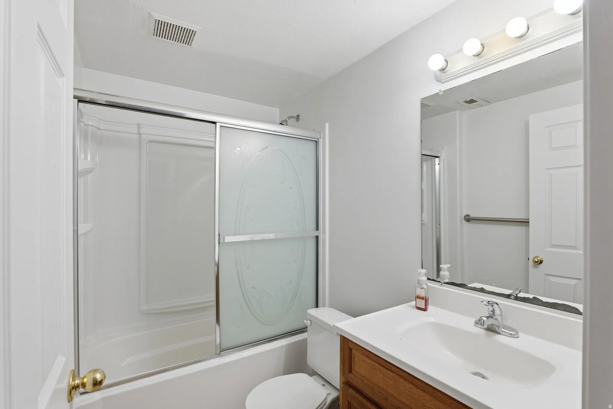 Bathroom featuring a tub/shower combination with sliding frosted glass doors, a vanity with a white countertop and integrated sink, a large wall-mounted mirror, and a brass-finished door handle