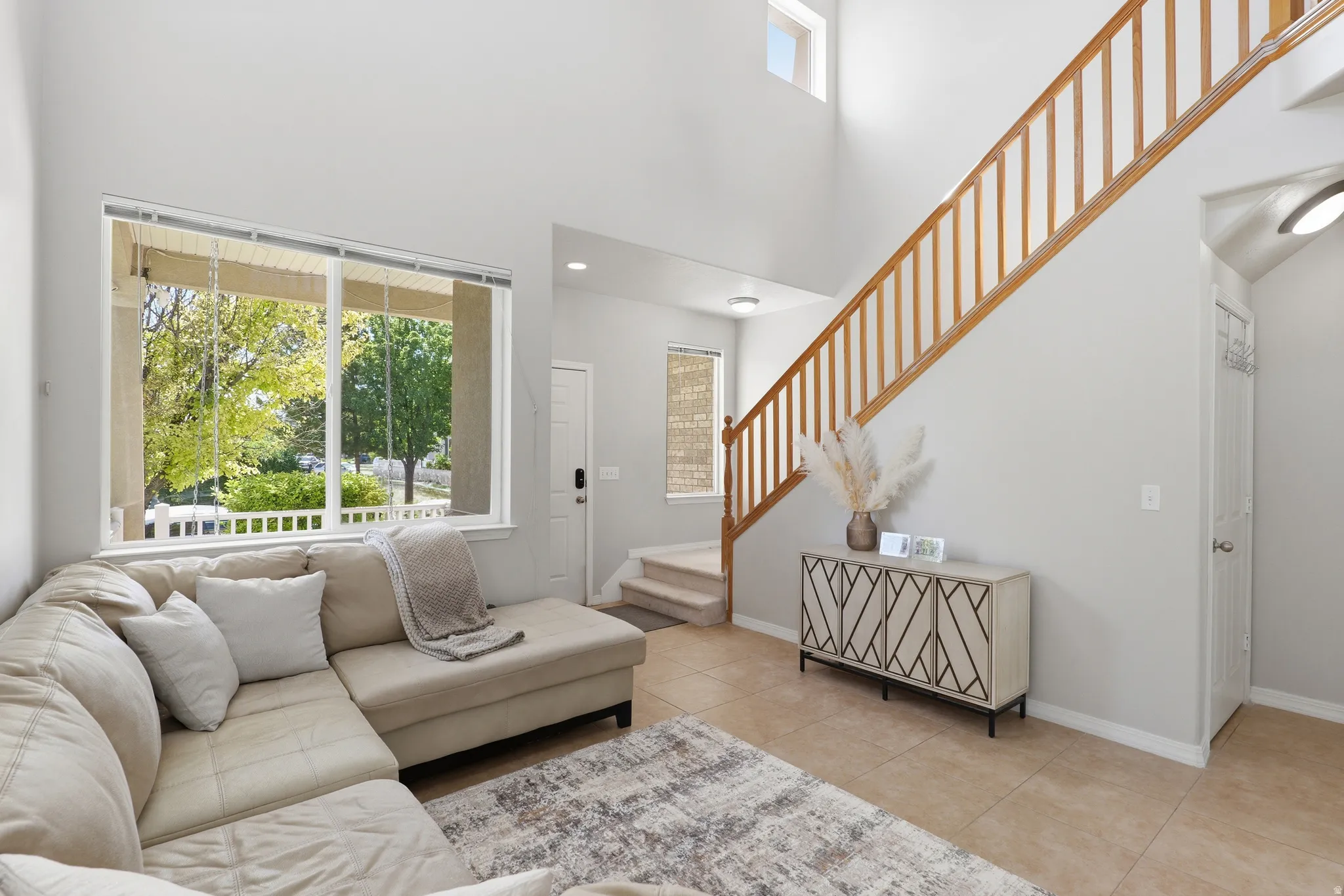 Two-story living area with high ceilings, large picture window with exterior views, wood-finish staircase with balusters, light tile flooring, and neutral wall tones