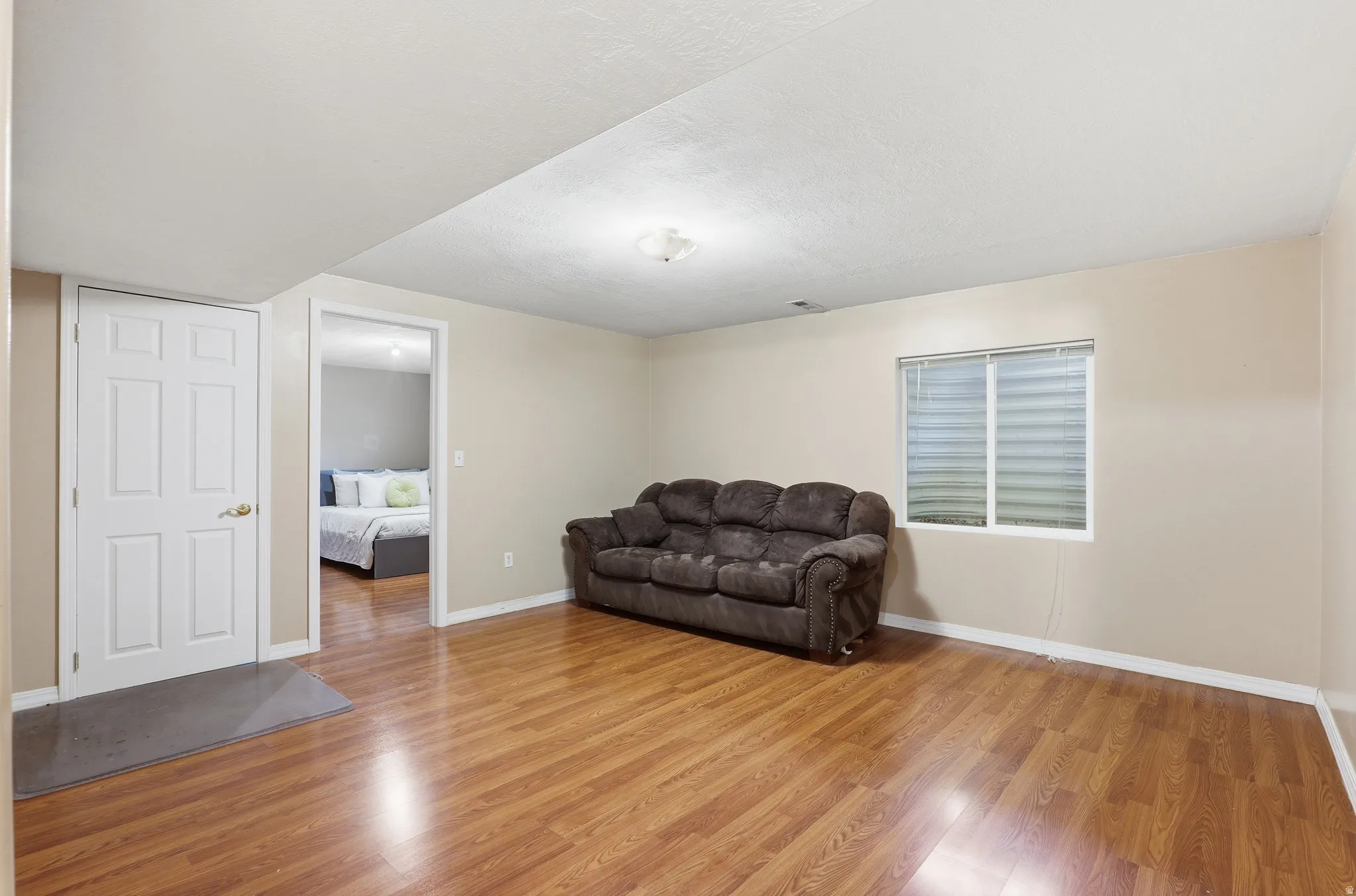 Spacious room featuring wood-finish flooring, a white window with horizontal blinds, and a white six-panel door with brass hardware