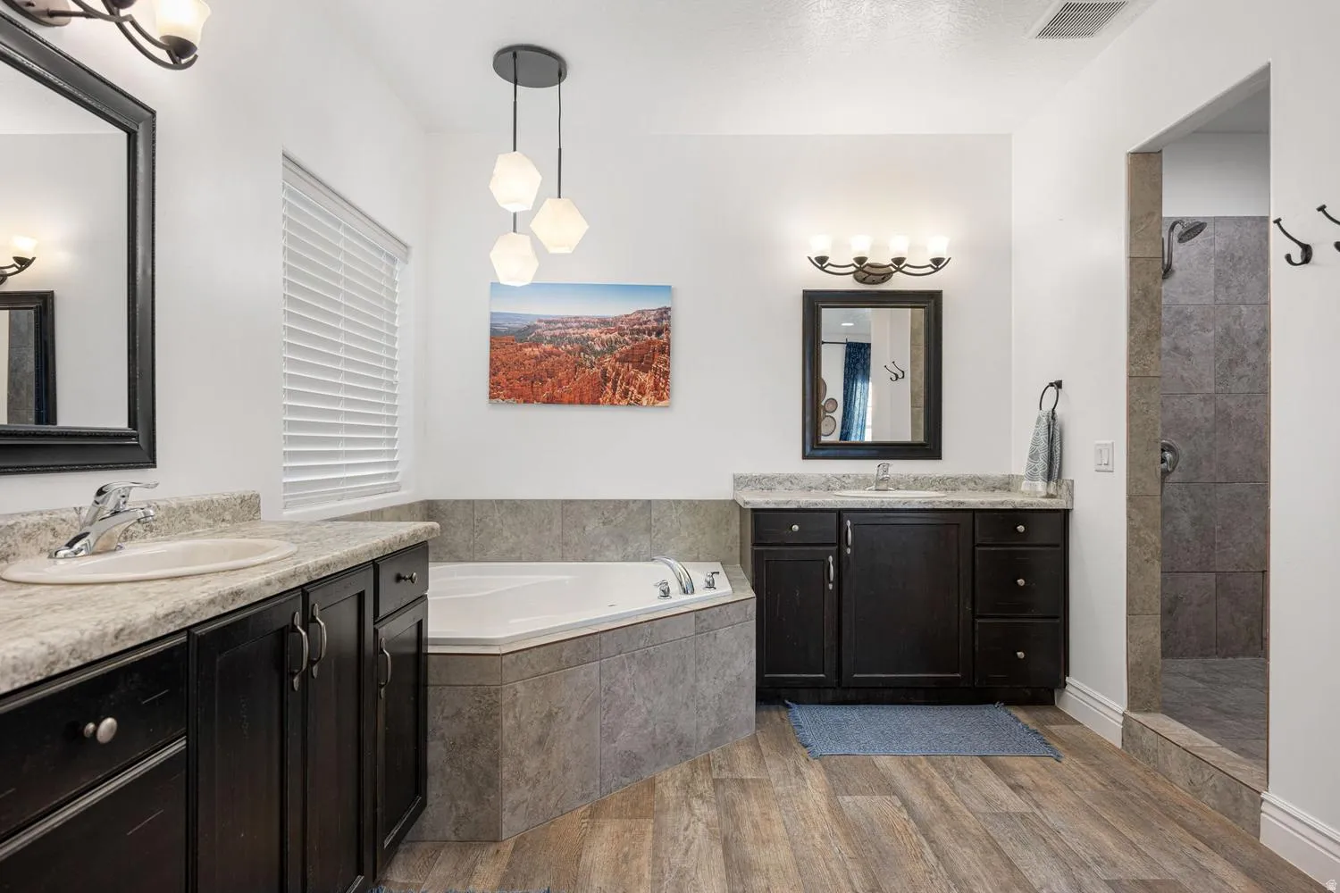 Corner soaking tub with gray tile surround, wood-finish flooring, dual dark wood vanities with light stone countertops and undermount sinks, and a walk-in shower with gray tile