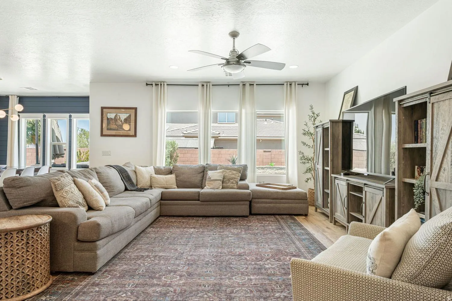 Spacious living area featuring a ceiling fan, wood-finish flooring, and multiple windows with roller shades