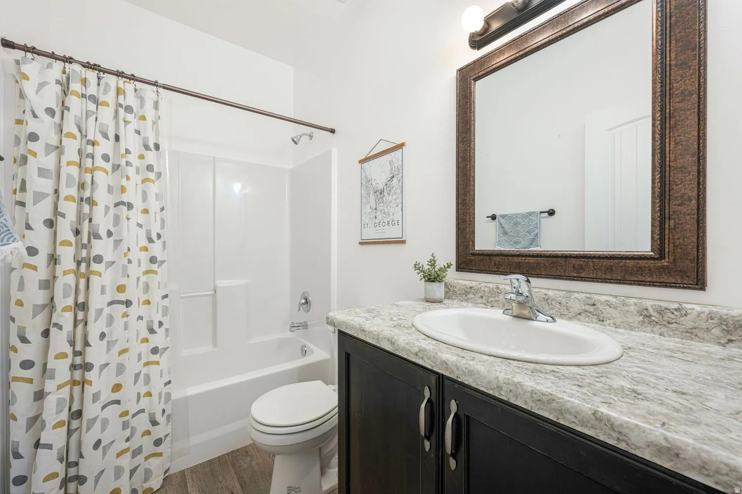 Bathroom featuring a white integrated tub and shower combination, a dark wood-finish vanity with a light speckled countertop, an oval undermount sink, and wood-finish flooring