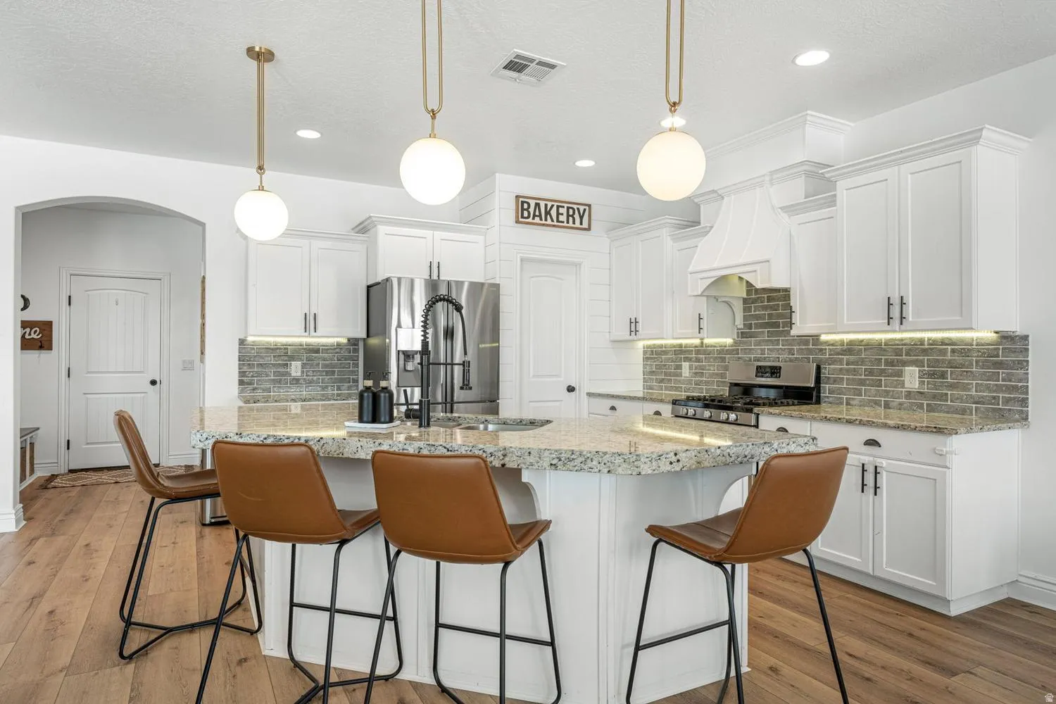 Kitchen featuring a large island with an integrated sink, light-toned granite countertops, white cabinetry with dark hardware, a gray brick-patterned backsplash, and wood-finish flooring