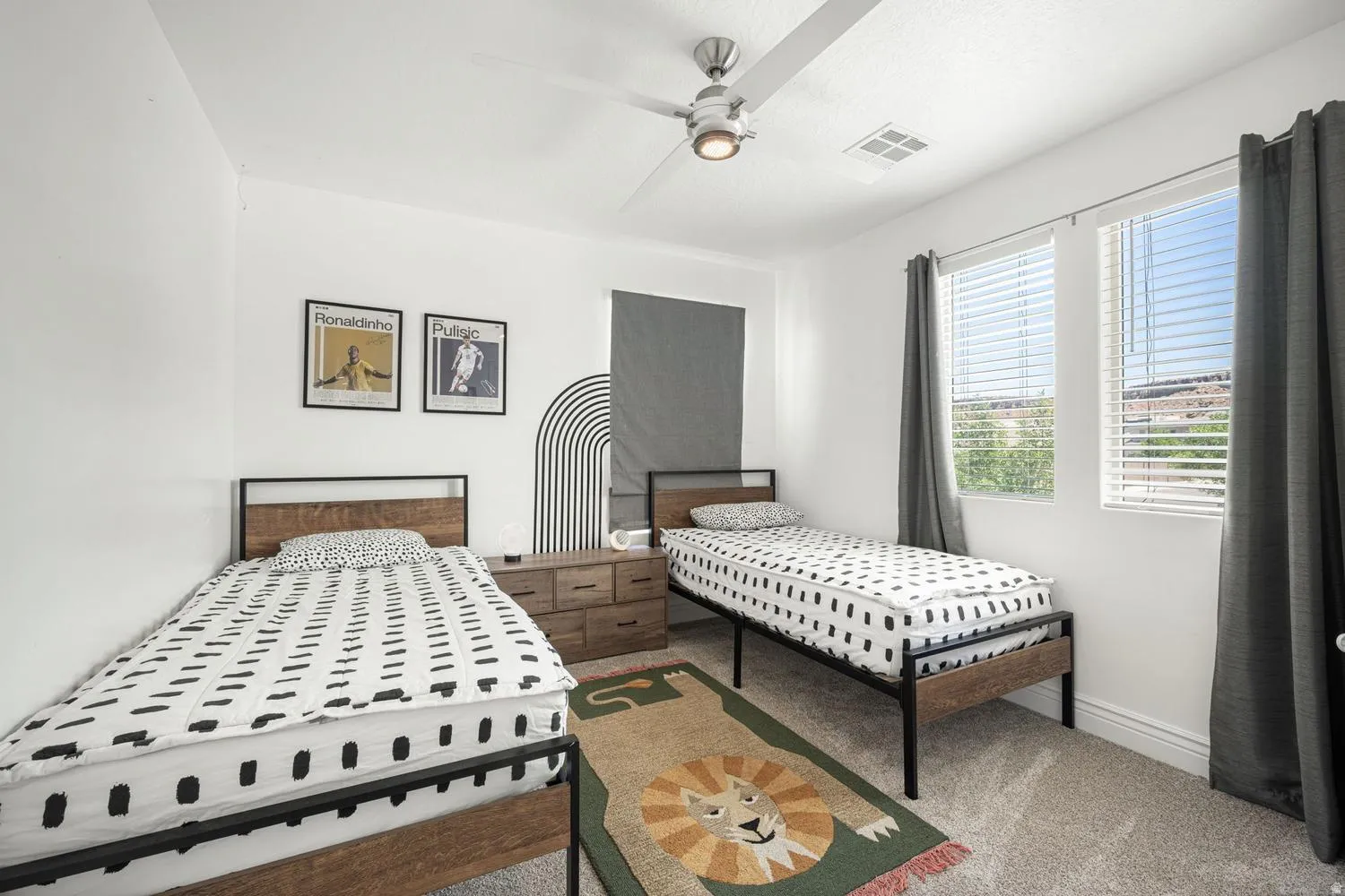 Spacious room featuring neutral wall tones, two windows with blinds and drapery, overhead ceiling fan with lighting, and plush carpeting