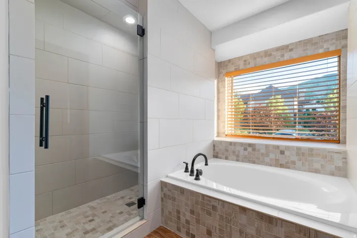 Primary bathroom featuring a built-in soaking tub with dark bronze fixtures, a glass-enclosed shower with subway tile, and a large window with wood blinds
