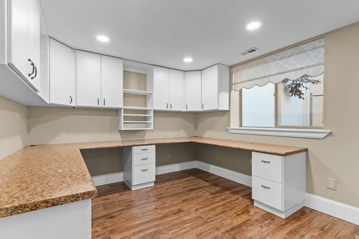 Dedicated workspace (office or craft room) featuring expansive built-in countertops, ample white cabinetry, wood-finish flooring, recessed lighting, and a window with natural light