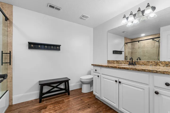 Bathroom featuring wood-finish flooring, a white vanity with a granite countertop, a shower with tiled walls and a glass enclosure, a built-in mirror, and a wall-mounted light fixture
