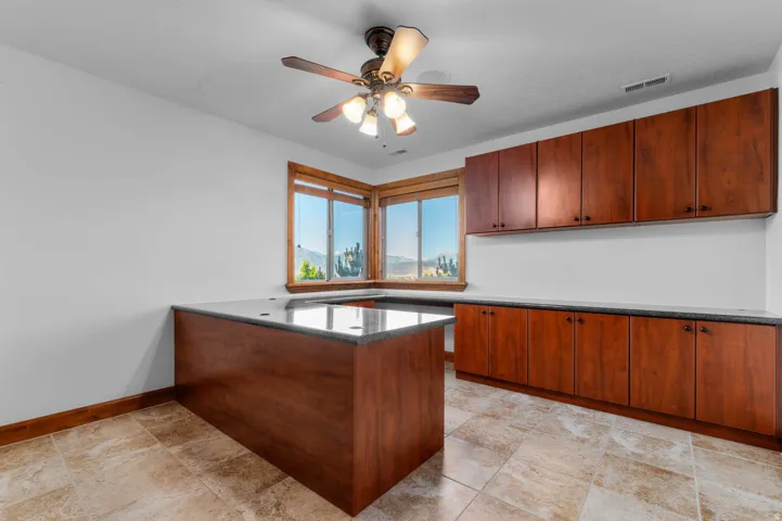 Spacious office or bedroom featuring extensive wood cabinetry with dark hardware, dark countertops, a ceiling fan with integrated lighting, and tile flooring