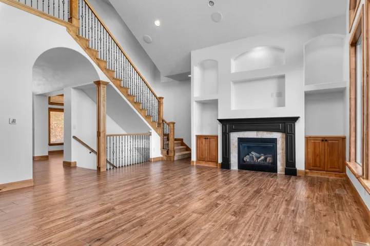 Expansive living space featuring wood-finish flooring, a grand staircase with wrought iron balusters, a fireplace with a black mantel and stone surround, integrated display niches, and recessed lighting