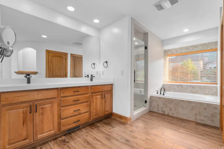 Primary bathroom featuring wood-finish flooring, a double vanity with white countertops, a large frameless mirror, a glass-enclosed shower, and a built-in bathtub with tiled surround