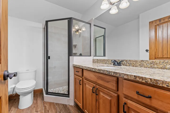 Bathroom featuring a glass-enclosed shower with pebble tile flooring and light-toned wall tiling, a vanity with a speckled countertop, integrated sink, and wood-finish cabinetry, a large wall-mounted mirror, and wood-finish flooring