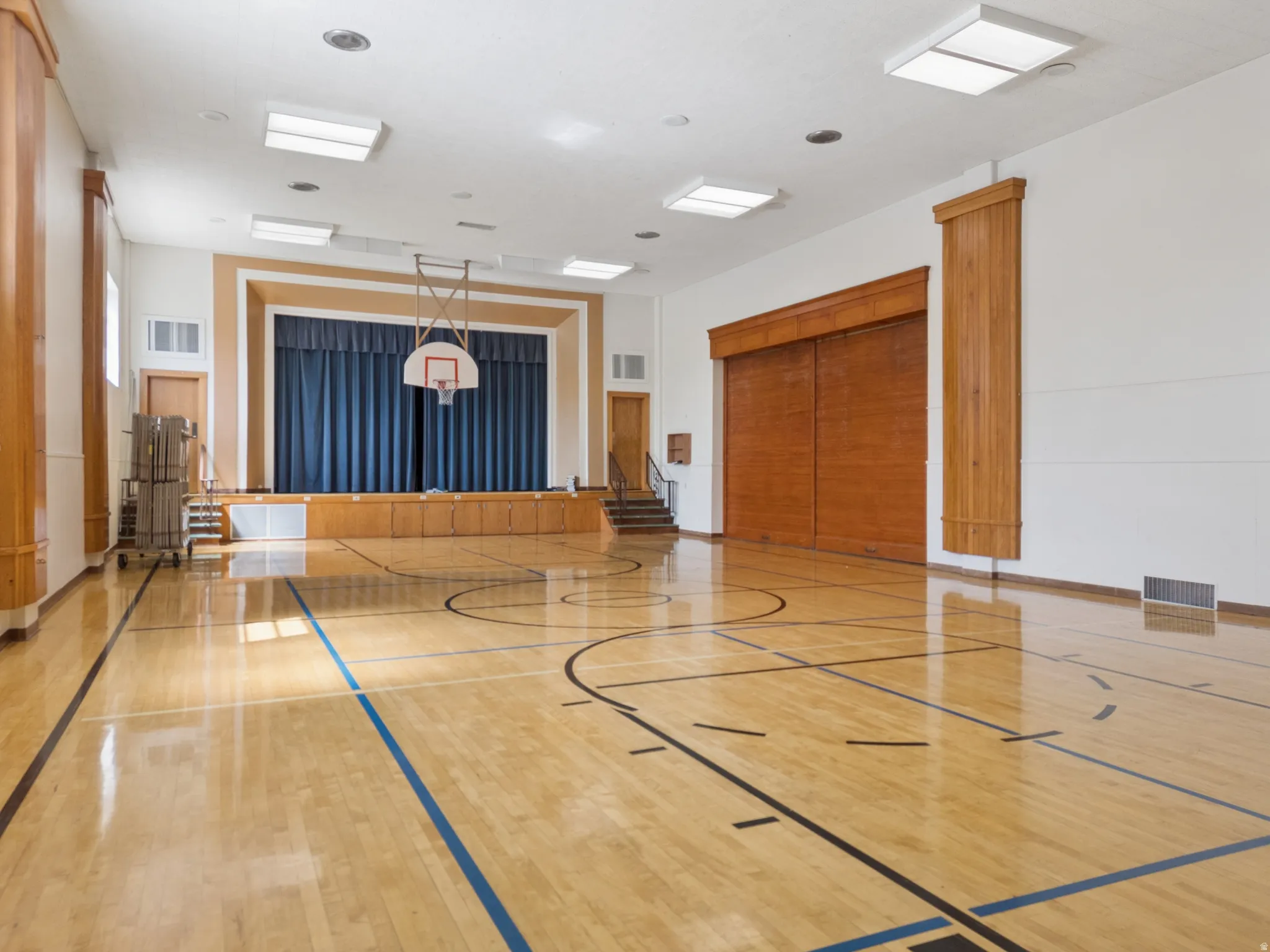 Expansive multipurpose room featuring polished wood flooring with court lines, a built-in stage with blue curtains, overhead lighting fixtures, and wood-paneled walls.