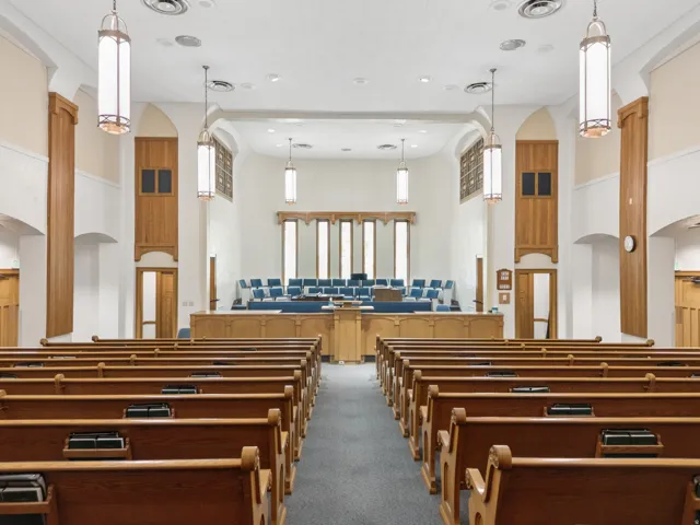 Spacious assembly hall featuring numerous wooden pews, a carpeted aisle, and a raised platform.