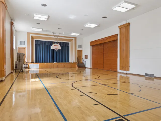 Expansive multipurpose room featuring polished wood flooring with court lines, a built-in stage with blue curtains, overhead lighting fixtures, and wood-paneled walls.
