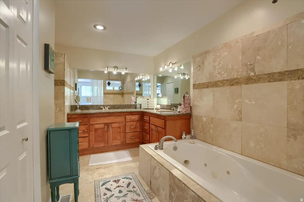 Bathroom featuring a jetted tub with stone tile surround, a corner vanity with wood cabinetry and stone countertops, dual undermount sinks, and a large wall mirror with overhead lighting