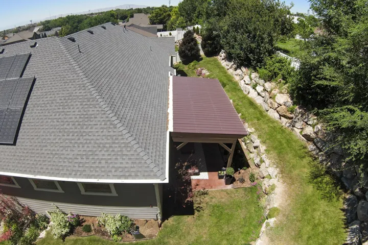 Exterior showcasing a gray shingle roof with integrated solar panels, a brown-roofed covered patio, a rock retaining wall, and established landscaping