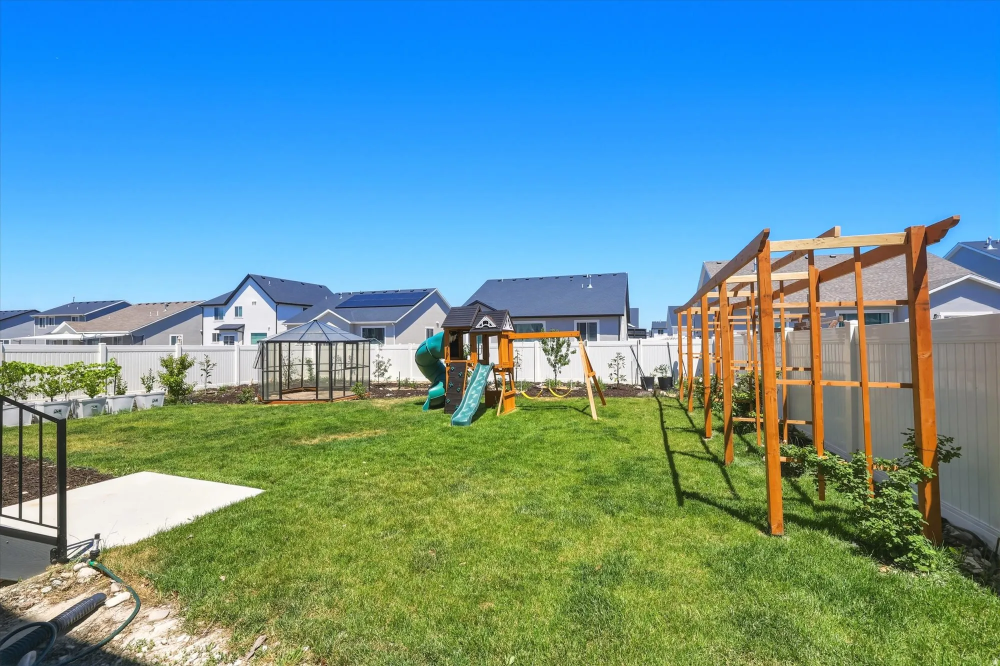 Expansive green lawn backyard featuring a white vinyl fence, a clear glass greenhouse structure, a natural wood arbor, and raised planter boxes