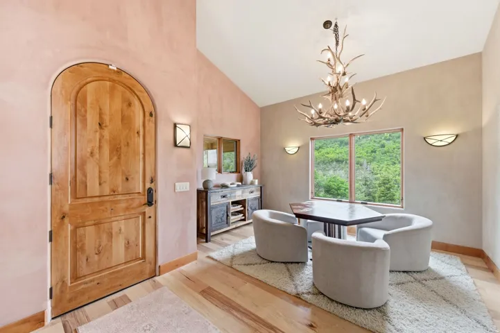 Arched wood-panel entry door, wood-finish flooring, vaulted ceiling, antler-style chandelier, and large window overlooking a forested landscape