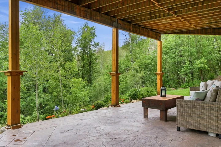 Covered patio featuring stamped concrete flooring, exposed wooden support beams, and a natural wooded backdrop