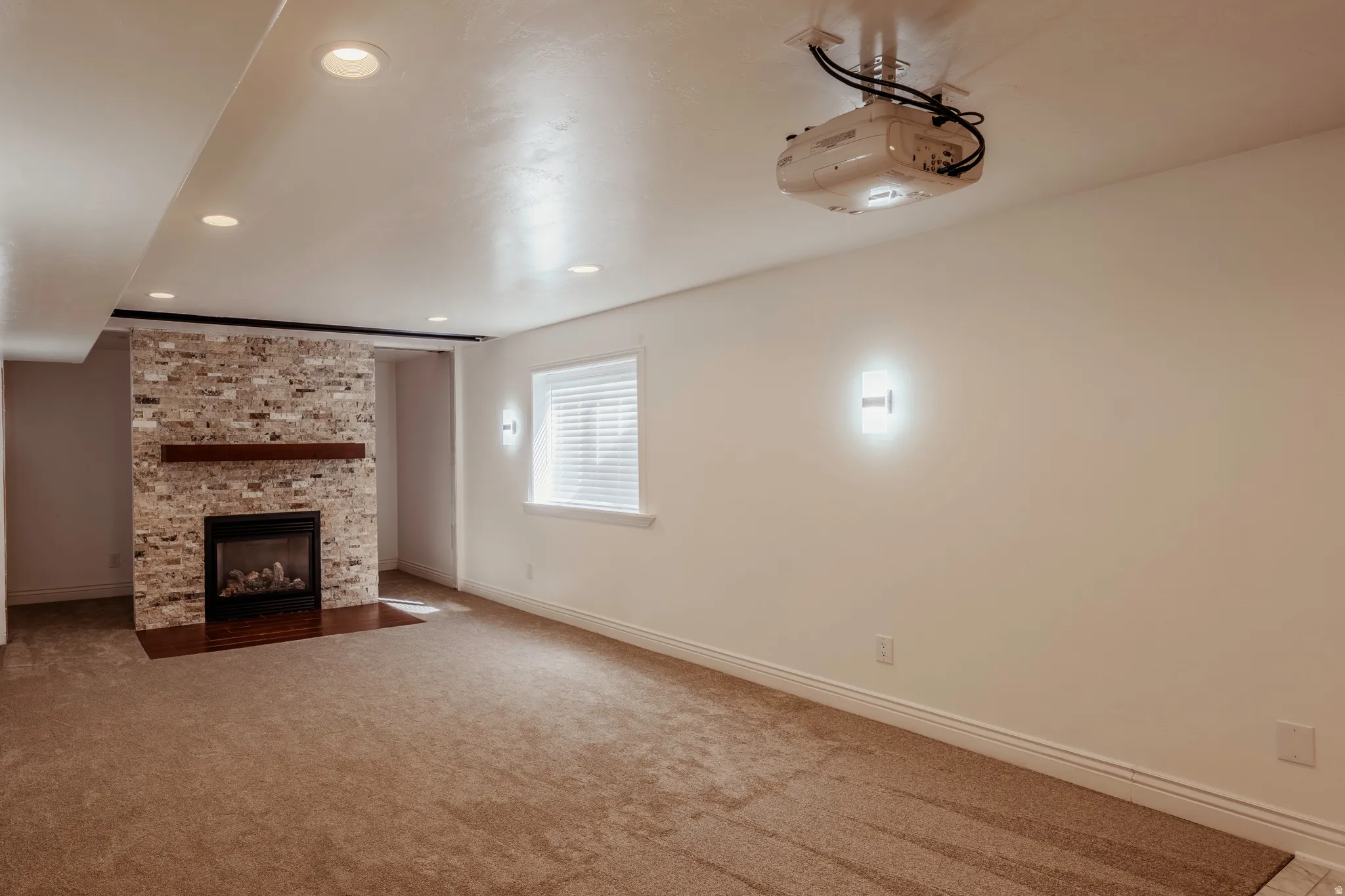 Carpeted room featuring a stone-clad fireplace with a wood mantel, a built-in projector, recessed lighting, and a window with blinds