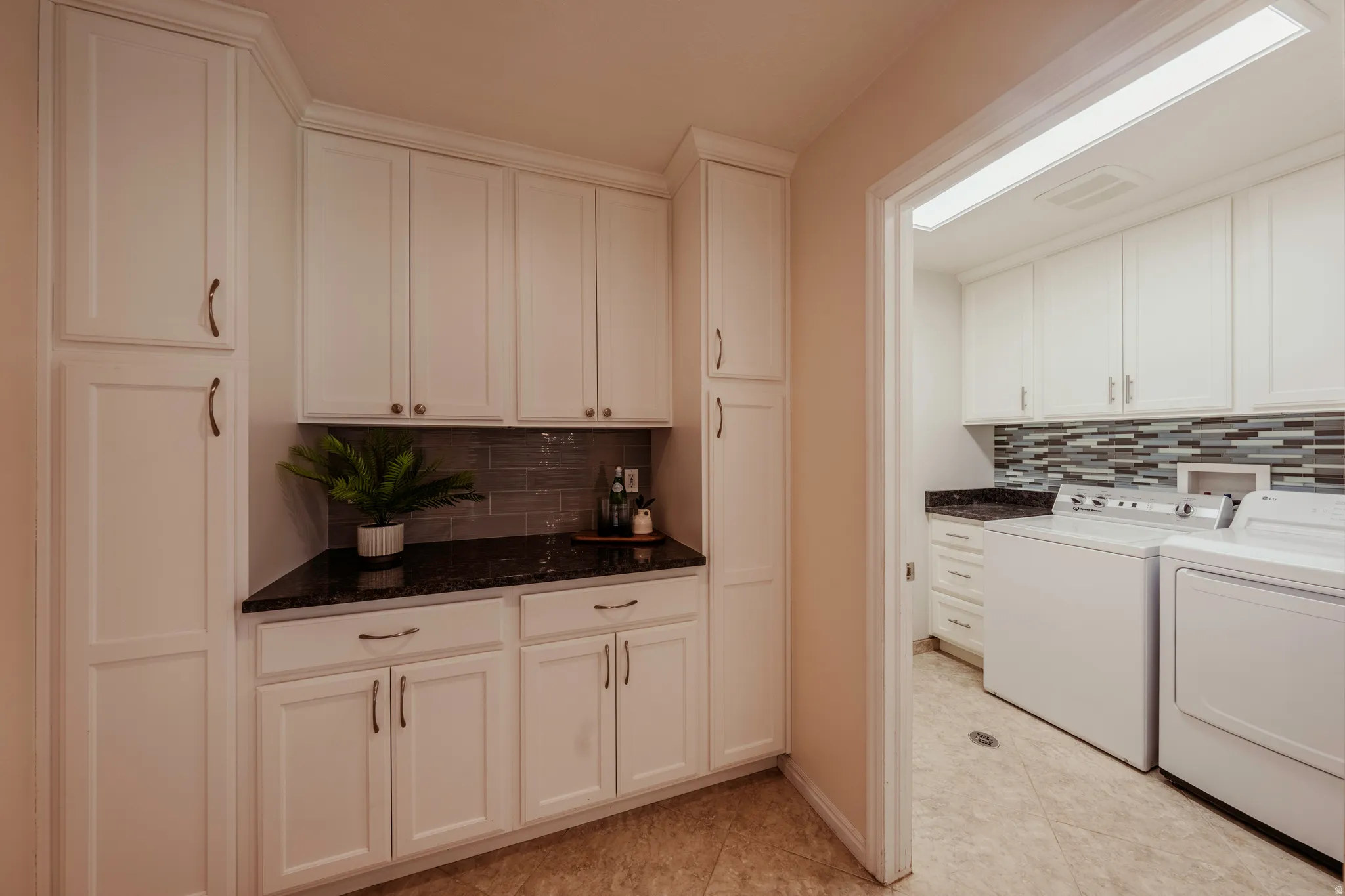 Functional utility room featuring extensive white cabinetry, dark countertop space, tile flooring, and a laundry area with a mosaic tile backsplash