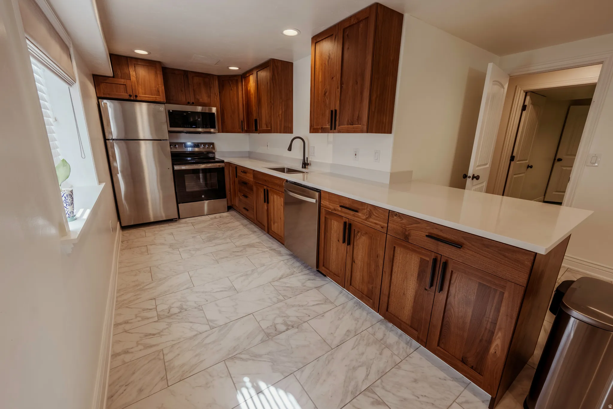 Contemporary kitchen featuring extensive wood cabinetry, white countertops, stainless steel appliances, a black gooseneck faucet, and light-toned, rectangular tile flooring