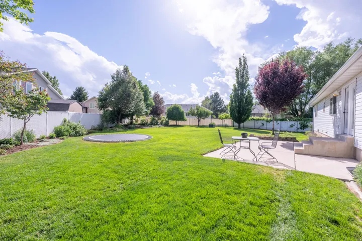 Expansive lawn area featuring a white vinyl privacy fence, mature trees, a concrete patio, and concrete steps leading to the house