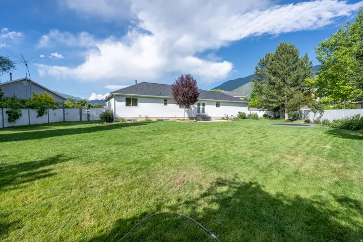 Expansive backyard featuring a broad lawn, mature trees, a white perimeter fence, and a patio area