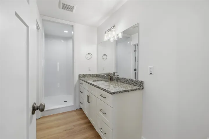 Bathroom featuring a walk-in shower with white surround, a vanity with a speckled countertop and integrated sink, a rectangular mirror, and wood-finish flooring