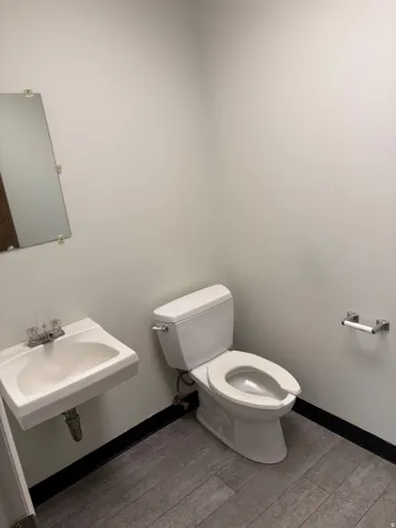 Compact restroom featuring a white wall-mounted sink, a two-piece toilet, a mounted mirror, and grey wood-finish flooring