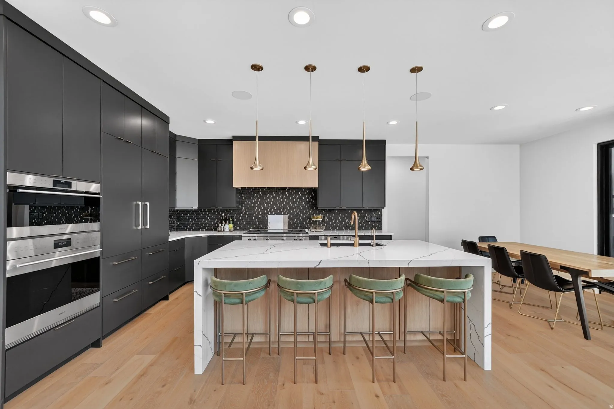 Spacious kitchen featuring an oversized waterfall island, light wood-finish flooring, dark cabinetry, built-in stainless steel appliances, and a mosaic tile backsplash