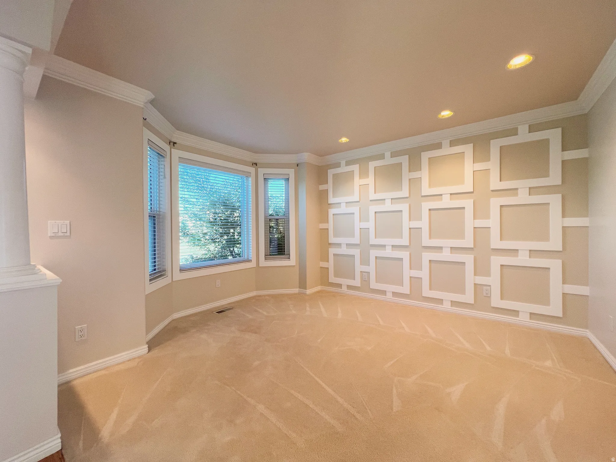 Spacious room featuring a decorative accent wall with geometric paneling, a bay window with blinds, recessed lighting, crown molding, and neutral-toned carpeting