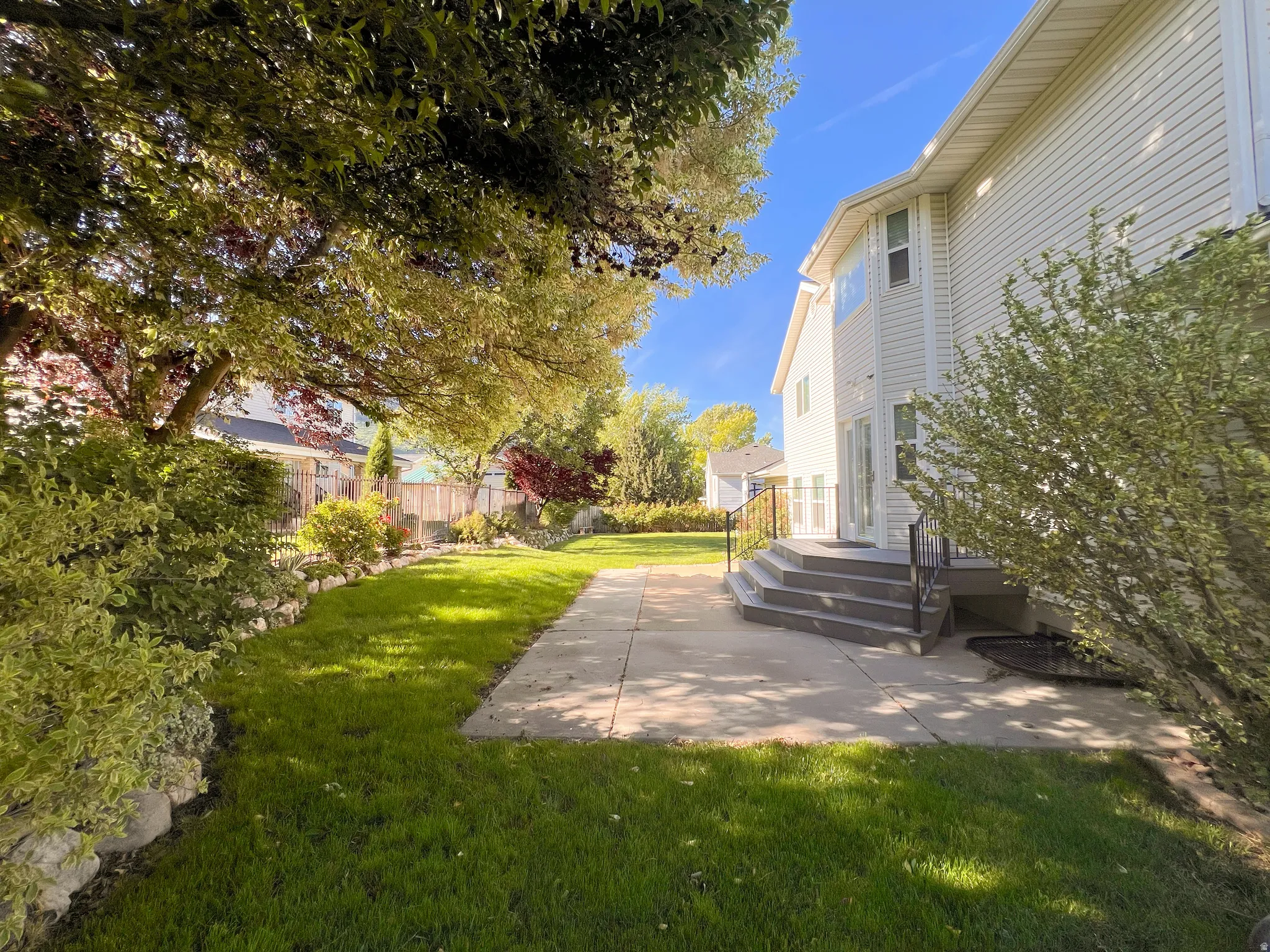 Rear exterior featuring a concrete patio, multi-tiered steps with black metal railings, and a expansive lawn with mature trees and shrubbery