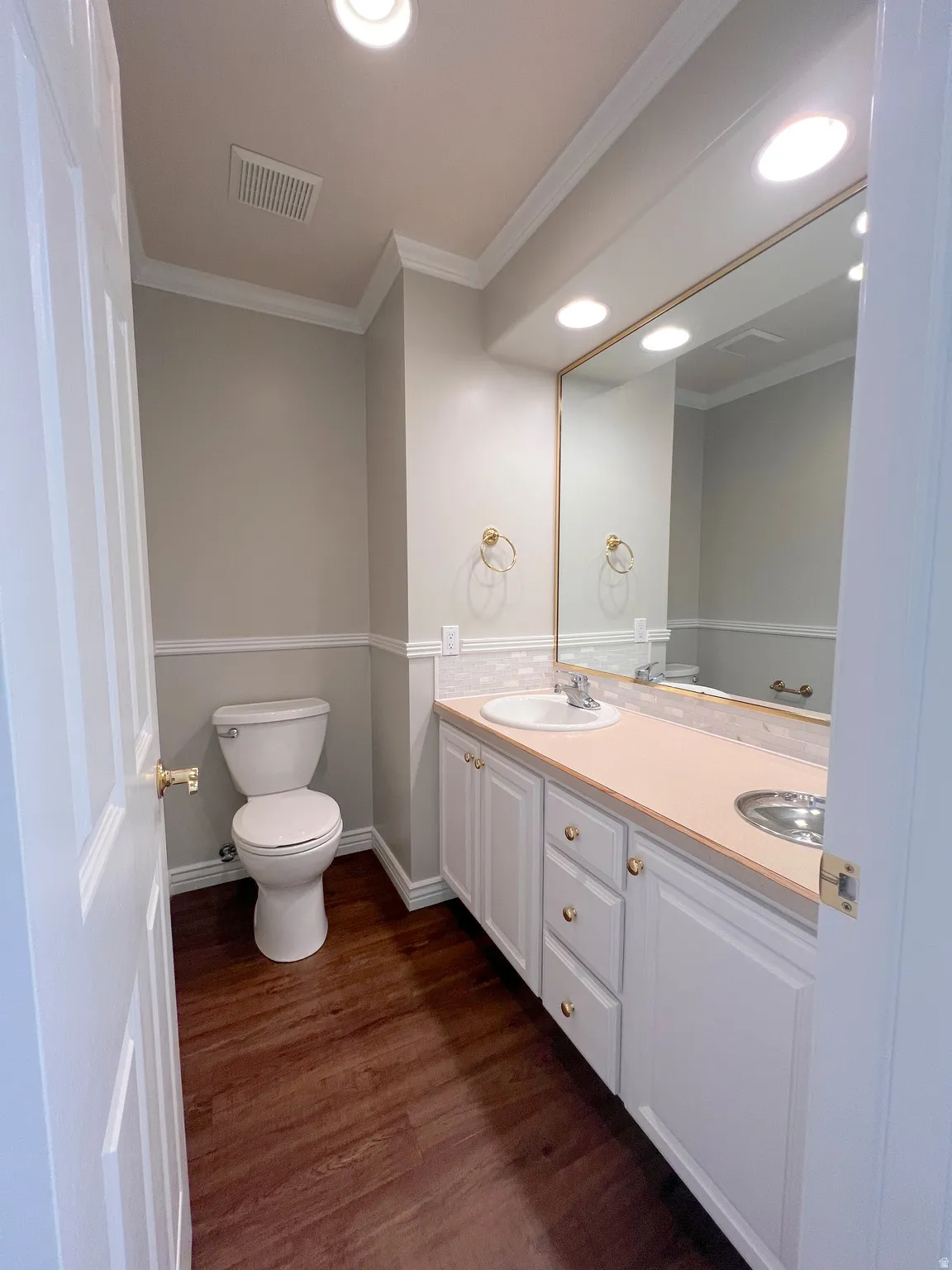 Bathroom with wood-finish flooring, a white vanity featuring a single sink and brass-toned hardware, a full-width wall mirror, and recessed ceiling lighting