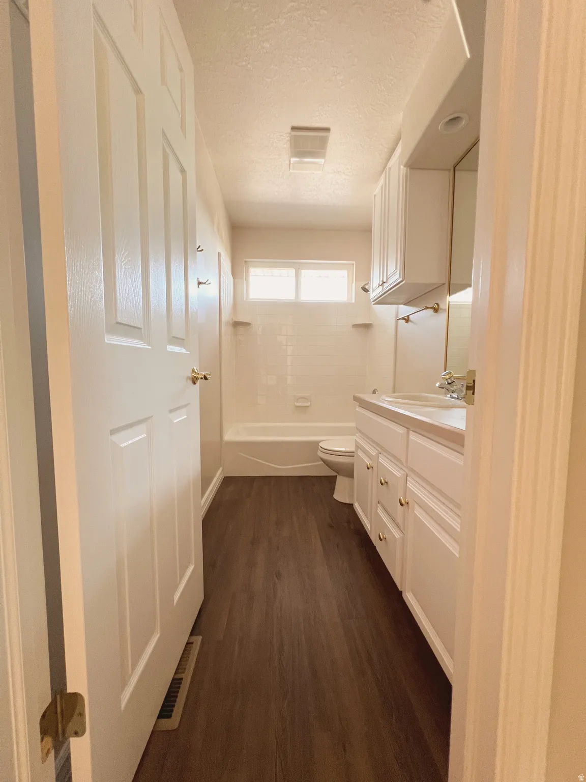 Bathroom featuring wood-finish flooring, a white vanity with gold-tone hardware, a built-in tub with overhead window, and white cabinetry