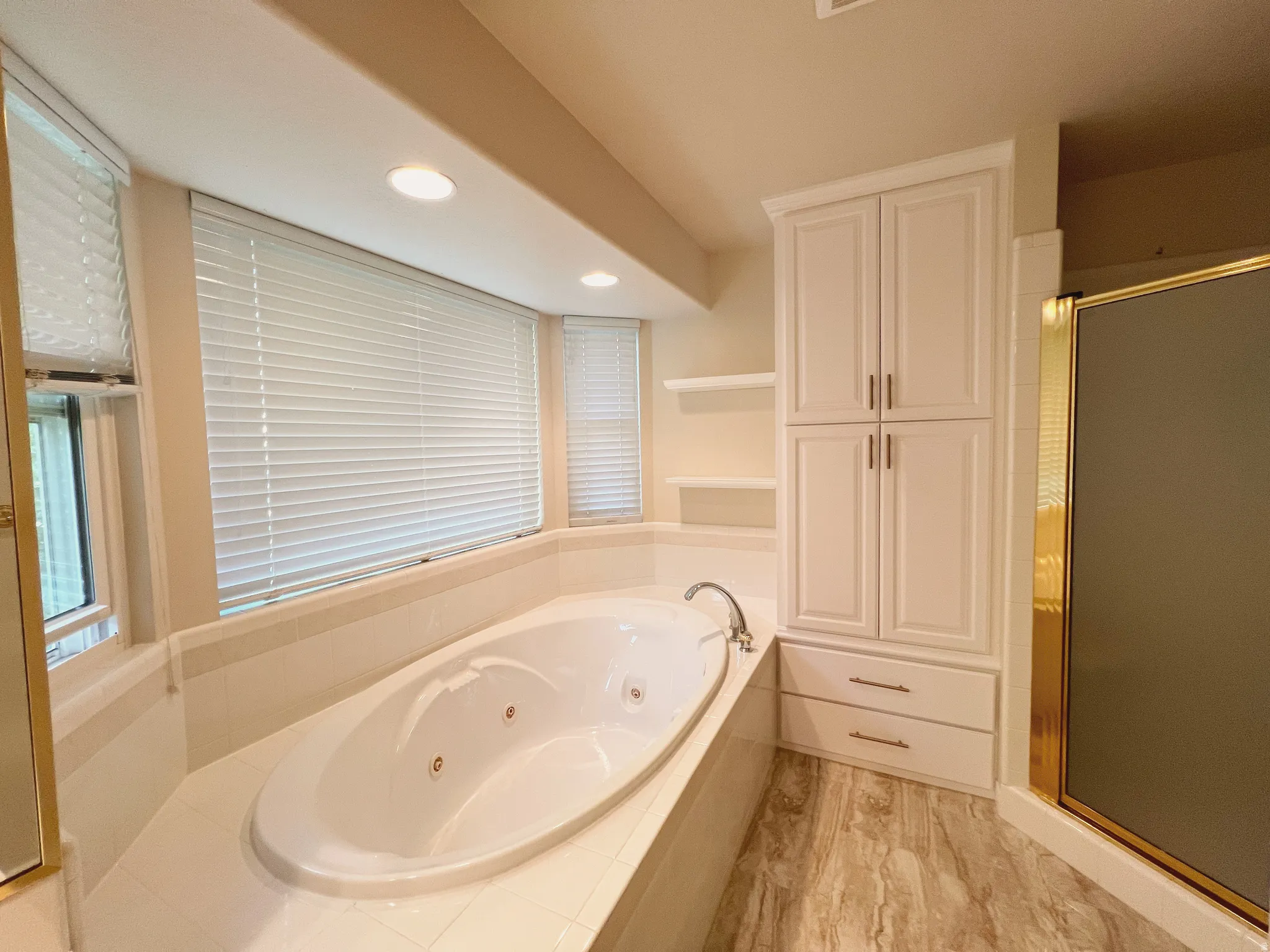Recessed jetted tub with tile surround, integrated built-in cabinetry with drawers, separate shower enclosure with gold-tone framing, and wood-finish flooring