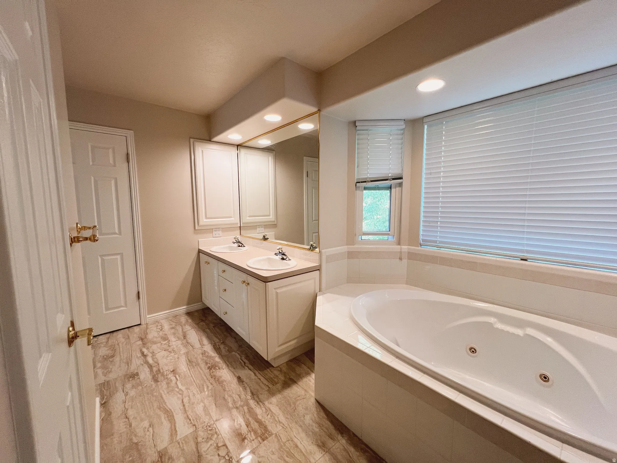 Corner jetted tub with tile surround, double vanity with integrated sinks, wood-finish flooring, and two windows with blinds