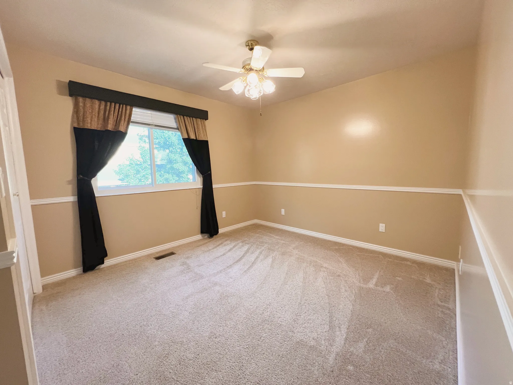 Carpeted room featuring a ceiling fan, a window with exterior views, and neutral-toned walls with chair rail molding