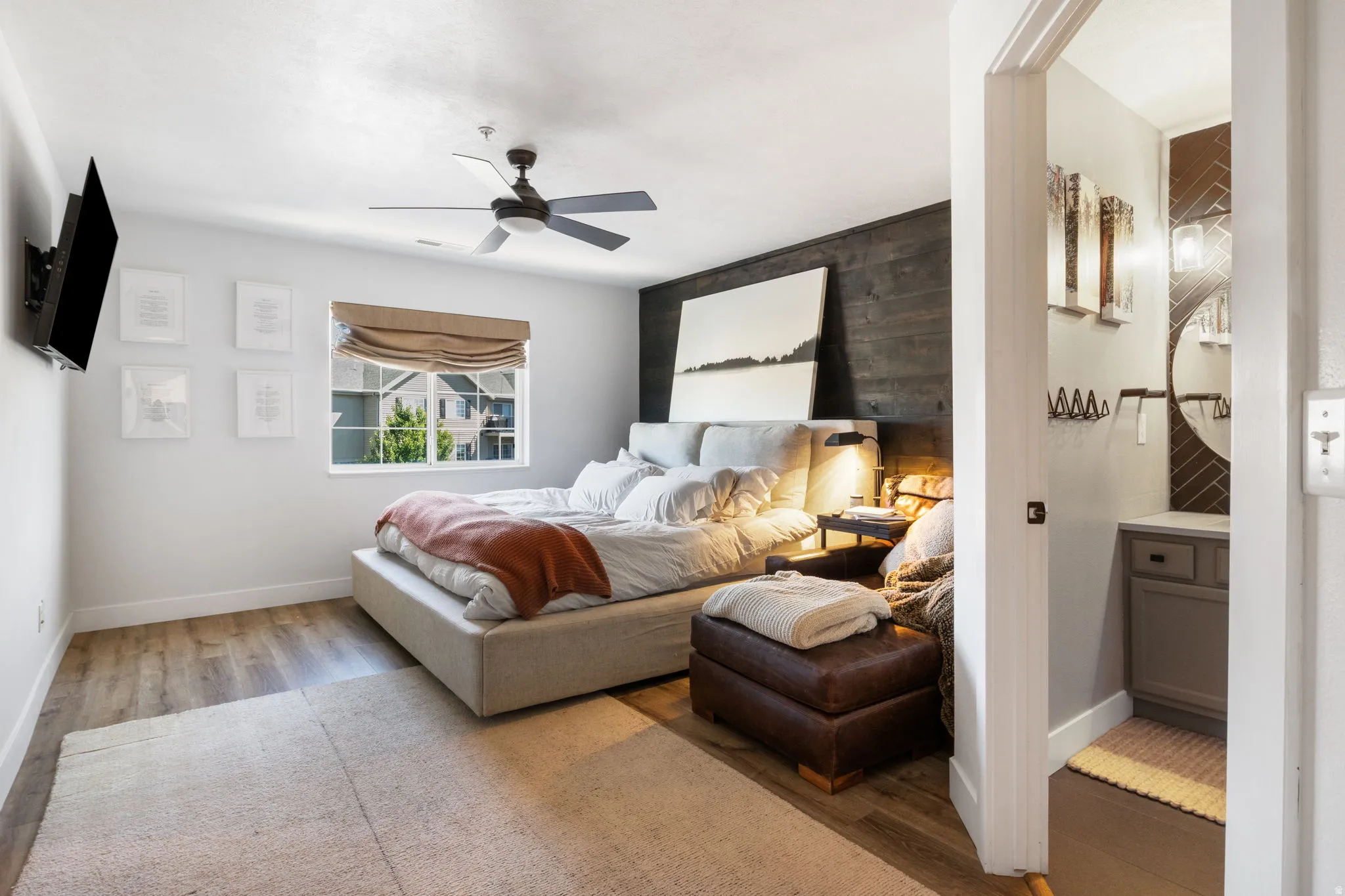Bedroom featuring wood-finish flooring, a ceiling fan with integrated lighting, and a window with a Roman shade