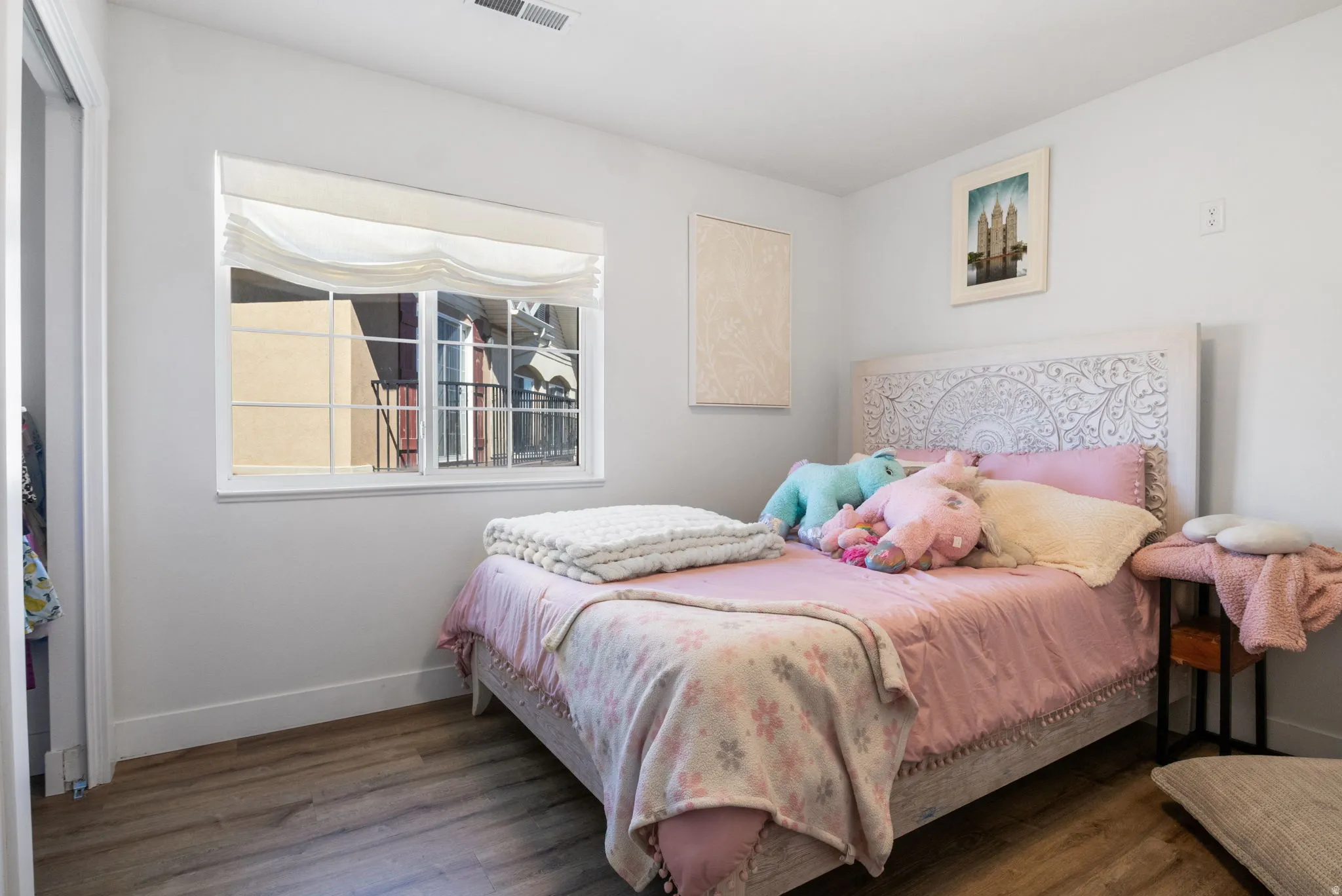 Bright room featuring wood-finish flooring, a white-framed window with a roman shade, and a carved wooden headboard