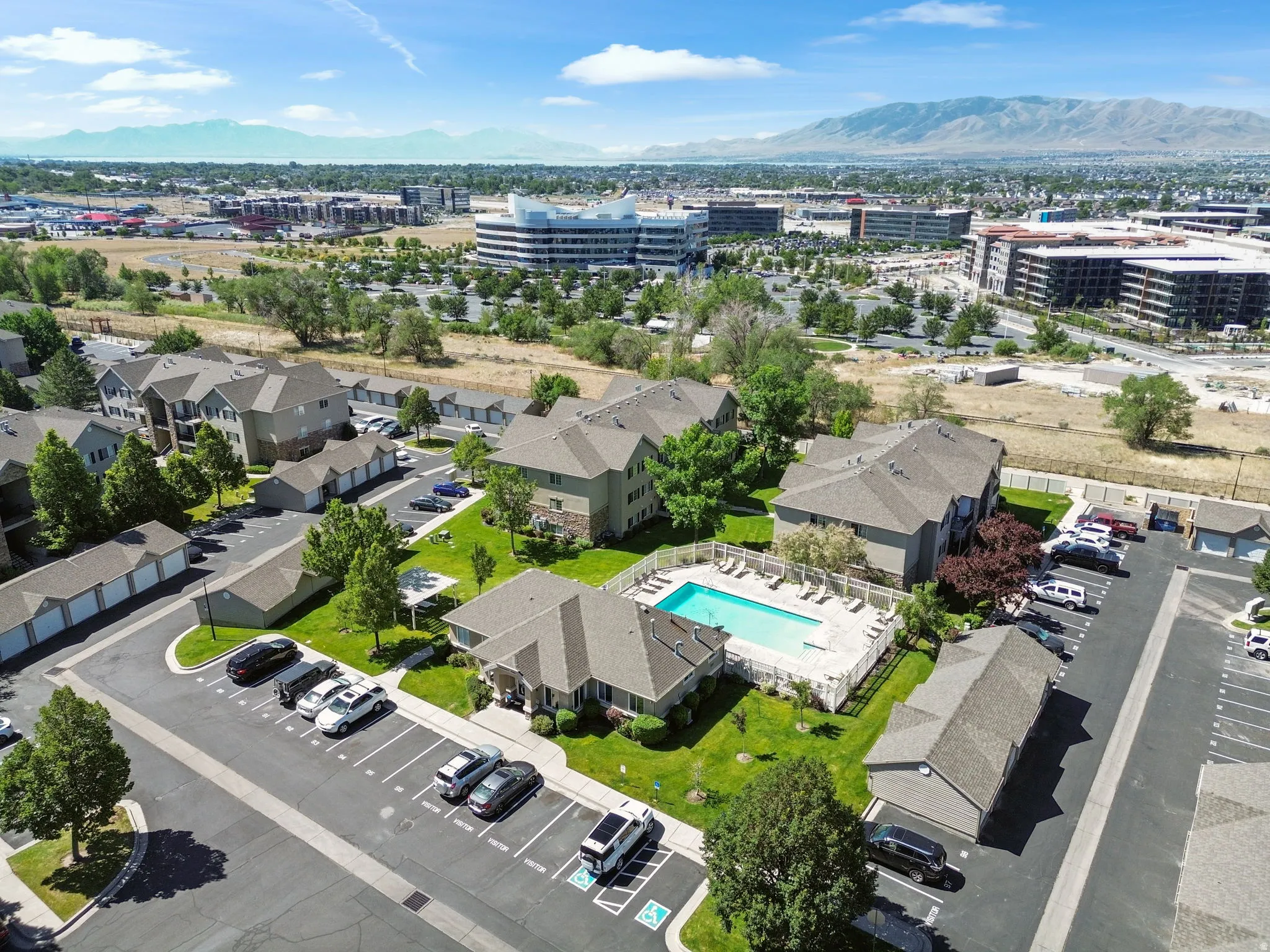 Community grounds featuring multiple residential buildings, a rectangular swimming pool with surrounding lounge chairs, paved parking areas, and detached garages