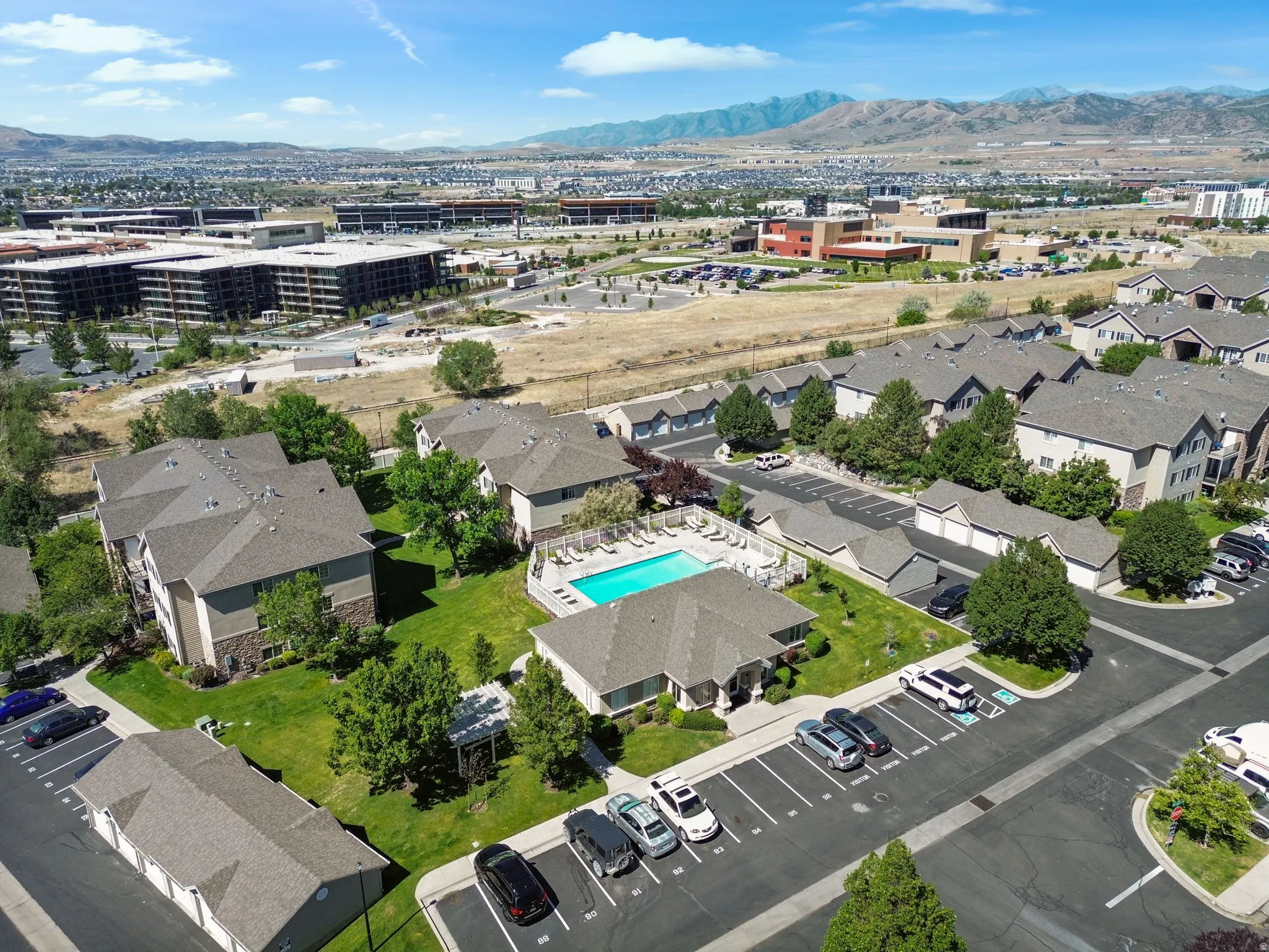 Expansive aerial view showcasing residential buildings with gray shingle roofs, a central swimming pool with surrounding patio, and dedicated parking areas